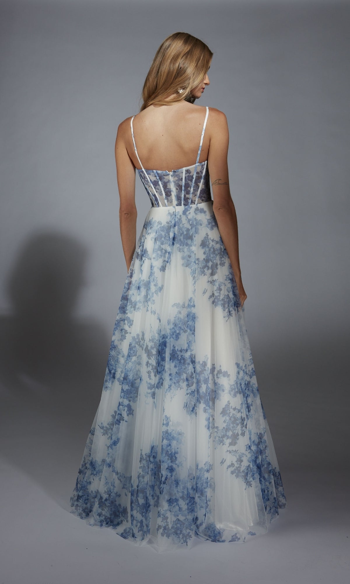 A woman in the Alyce 61752 long prom dress, a sleeveless, floor-length white gown with blue floral prints and a corset bodice, stands with her back to the camera against a plain gray background.